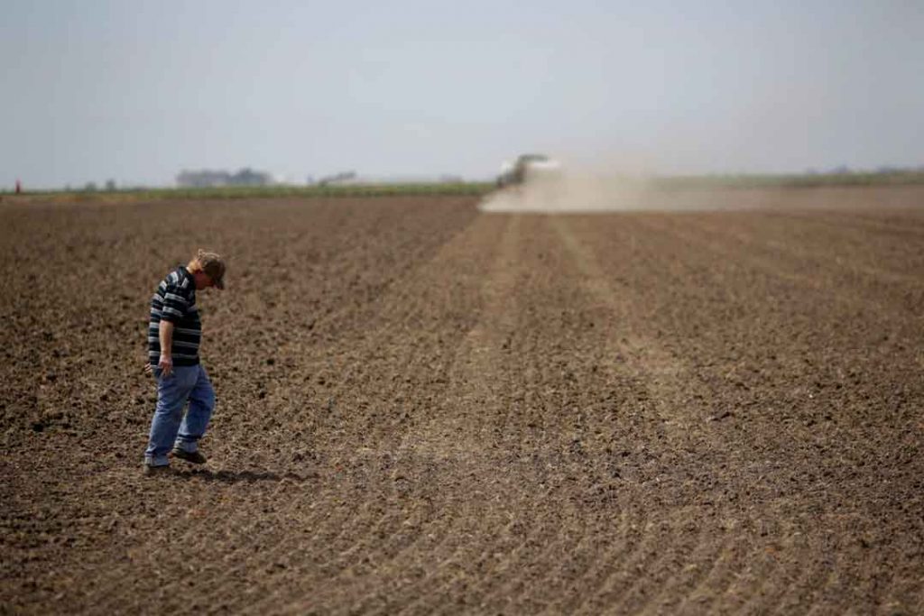 A man walking across a plowed dirt field with a plow in the background